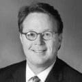 A black-and-white portrait of a man with short, neatly combed hair, wearing glasses, a suit jacket, a dress shirt, and a striped tie. He is smiling and looking directly at the camera, with a neutral background.