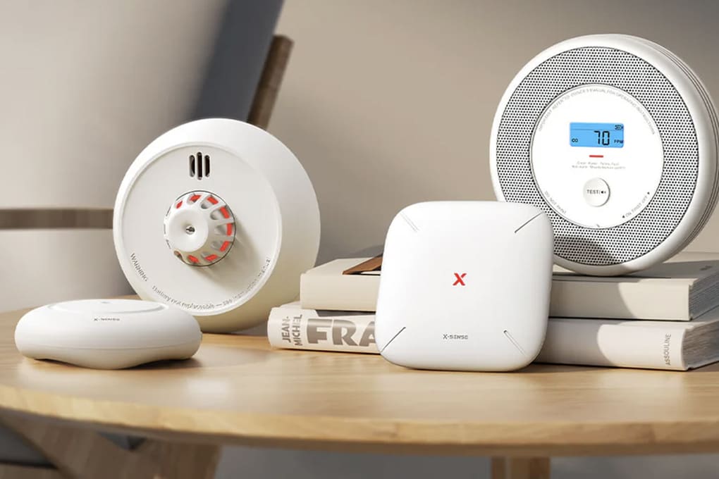 Smart home devices on a wooden table, including a smart smoke detector with a digital display and a white router
